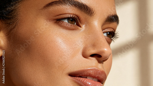 Close-up portrait of a woman with glowing, dewy skin and natural makeup. Features focused on eyes, brows, and glossy lips under warm sunlight.