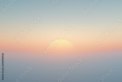 Soft sunrise emerging above misty horizon, blending gentle blue tones with warm pink, orange, and yellow hues, creating a calm minimalistic sky scene of ethereal beauty.