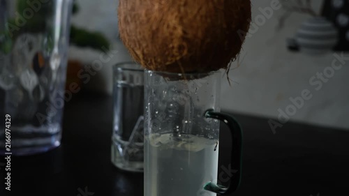 Fresh coconut water dripping from a coconut into a glass mug on a dark kitchen counter, with a soft blurred background.