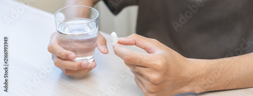 Bild auf Leinwand Sick asian young man holding tablet pill on hand, painkiller medicine from fever, stomach pain, headache for treatment, take drug, vitamin with glass of water on table at home