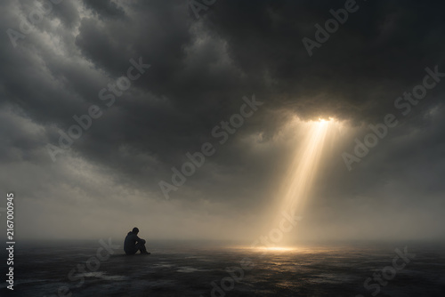 Small human sitting beneath an enormous dark cloud while a narrow beam of sunlight, depression, emotional heaviness and hope concept
