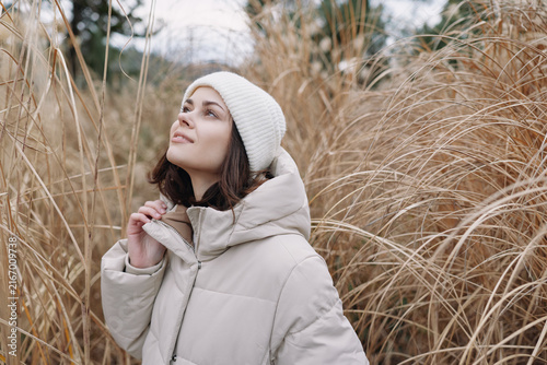 Fotografie Woman in a light winter coat and white beanie gazes upward among tall grasses, capturing a serene outdoor portrait in soft natural light