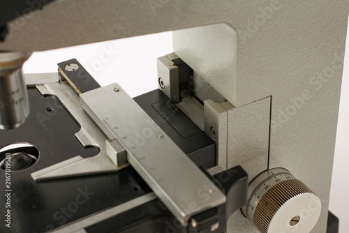 Detailed view of the stage assembly of an old scientific microscope, featuring a glass specimen slide, adjustable mechanical holder and graduated control mechanism.