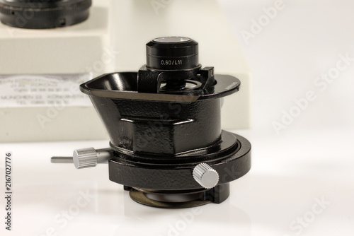 Close-up of a vintage microscope condenser with optical lens, adjustable aperture housing and precision control knobs, showing detailed black metal construction on a white surface.