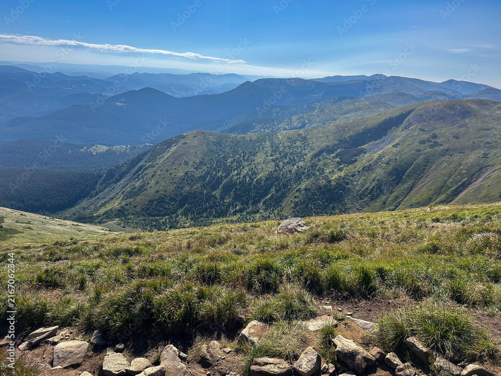 Fototapeta premium Wide alpine hiking trail descends over a vast mountain valley with blue sky and distant ridges, ideal for travel, trekking and outdoor adventure themes.