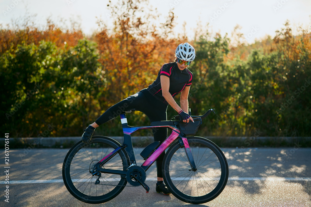 Fototapeta premium Female Cyclist Stretching On Aerodynamic Road Bike Before Training Ride In Autumn Sunrise