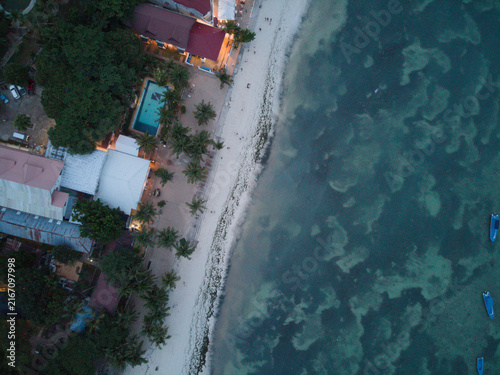 A straight-down drone frame shows darkening water, white sand, rooftops and a swimming pool.