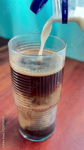 A close-up vertical shot showing milk being poured from a plastic bottle into a tall, ribbed glass of dark black coffee.