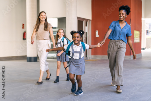 Mothers walking hand in hand with their happy multiethnic girls, wearing school uniforms and backpacks, on their first day back to school