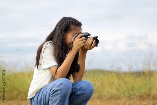 Young girl photographer squatting outdoors taking photos with a DSLR camera against sky background.