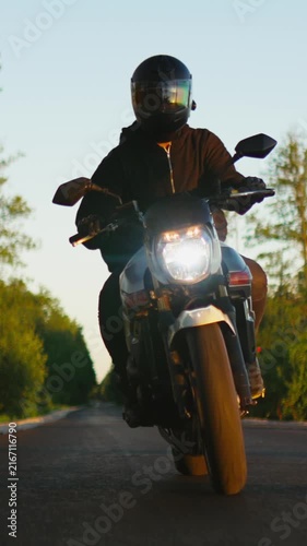 Lone motorcyclist in dark gear rides a powerful street bike along an empty rural road at golden hour, headlight shining brightly through the tranquil sunset landscape