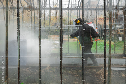 A man in a mask and overalls sprays a greenhouse with pesticides and insecticides against pests in autumn.