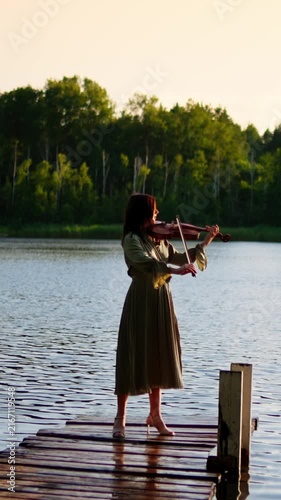 Young woman playing violin on a wooden lakeside pier at sunset, surrounded by calm water and lush green trees in a peaceful outdoor nature setting