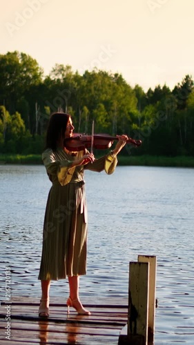 Elegant woman playing violin on wooden lakeside pier at golden sunset, surrounded by calm water and lush green forest in peaceful outdoor nature scene