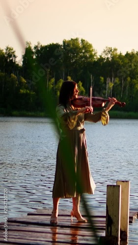 Woman playing violin on wooden lakeside pier at golden sunset, creating peaceful music in nature with calm water, soft light, and tranquil forest reflections