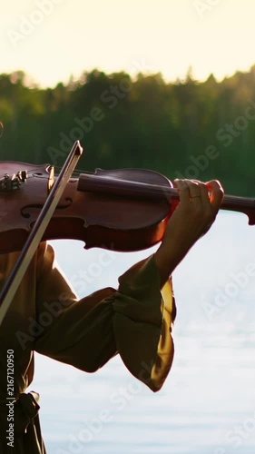 Violinist performing serene melody outdoors by tranquil lakeside at sunset, warm golden light illuminating graceful hands and strings in peaceful natural setting