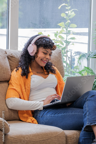 Smiling young woman with curly hair wearing pink headphones, working on laptop while sitting in armchair at home, cozy lifestyle and remote work concept
