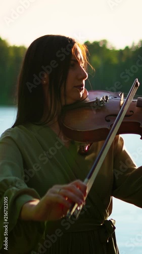 Serene lakeside violin performance at sunset as a focused woman in flowing dress plays a soulful melody in warm golden light against a calm water backdrop
