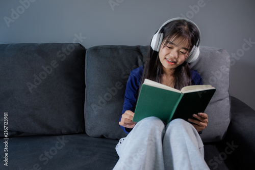 Woman is wearing headphones and reading a book while relaxing on the sofa in the living room