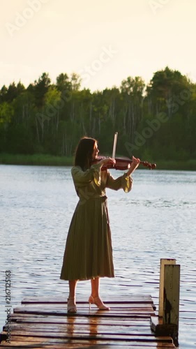 Young woman playing violin on rustic wooden pier at tranquil lakeside during golden sunset, creating serene, reflective atmosphere against forested shoreline