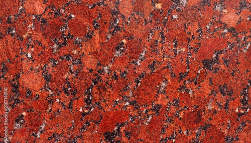 Polished red granite stone surface texture with scattered black biotite mineral speckles and white quartz crystal inclusions