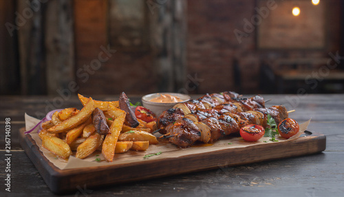 Grilled meat skewers with crispy potato wedges cherry tomatoes and dipping sauce on rustic wooden board warm restaurant lighting