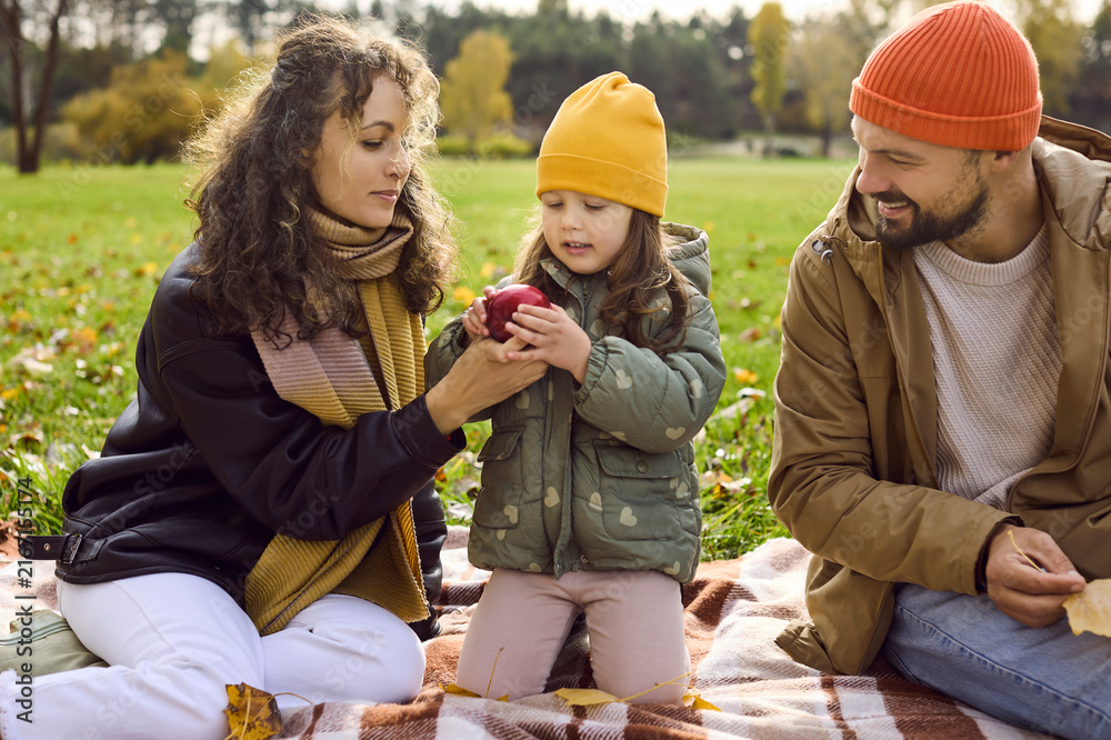Fototapeta premium Happy family in autumn park, smiling daughter. Parents share an apple with their child on a picnic blanket. Warm colors and scarves show love and bonding. Family togetherness and seasonal joy.