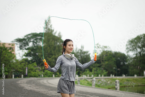 Upper body close-up of a cheerful Asian woman smiling while swinging a colorful jump rope during a fitness workout near a lake.
