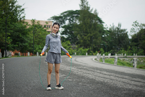 Full length photo of a smiling Asian woman standing on a road holding a skipping rope, ready for a jump rope exercise session outdoors in a park.