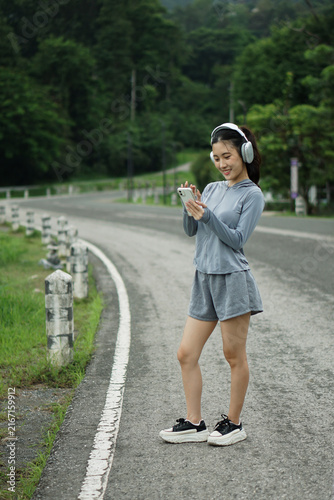Full length portrait of a young Asian woman in fitness wear checking her smartphone on a scenic road near green mountains.