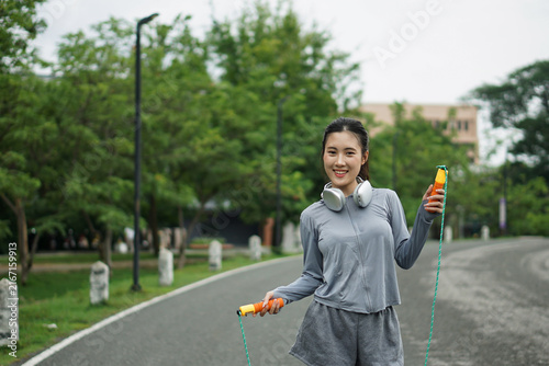 Full length photo of a smiling young Asian woman standing on a road holding a skipping rope, preparing for a cardio workout outdoors.