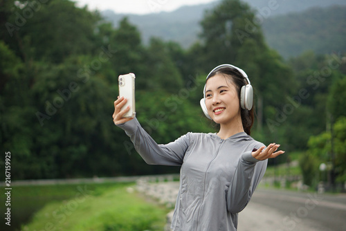 Upper body shot of a smiling Asian woman holding her smartphone to take a selfie or video call while gesturing happily outdoors.