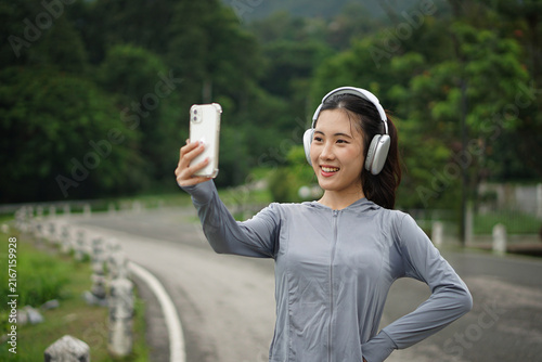 A happy Asian woman in activewear taking a photo with her smartphone while wearing headphones during an outdoor fitness session.