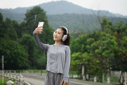An Asian woman wearing headphones and gray activewear looking up happily as she holds her mobile phone high to take a portrait shot outdoors.