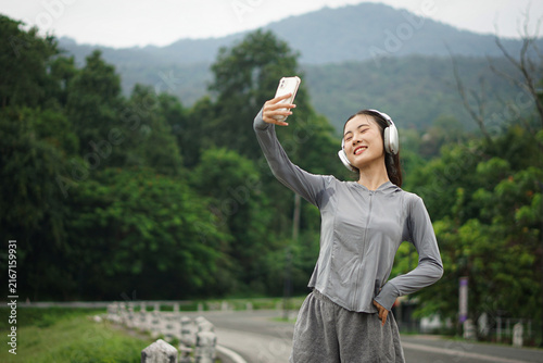 A peaceful young Asian woman closing her eyes with a happy smile while holding up her phone for a selfie against a backdrop of mountain greenery.