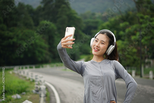 A charming Asian woman tilting her head with a warm smile while taking a selfie with one hand on her hip during an outdoor fitness session.