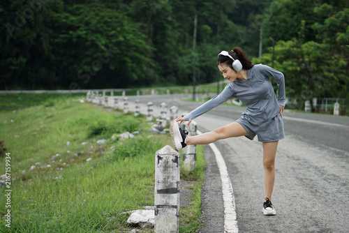 Full length photo of an Asian woman in fitness gear stretching her hamstring on a concrete post along a road near green park scenery.