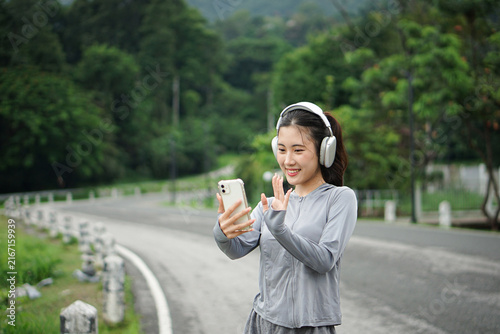 A friendly Asian woman waving her hand while having a video call on her smartphone in a scenic outdoor park environment.