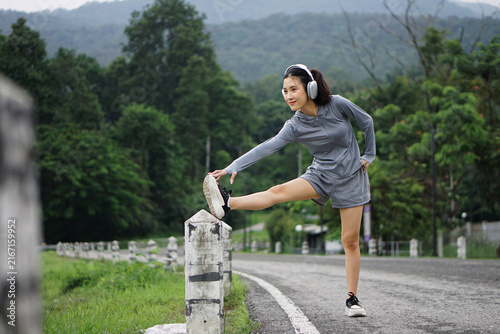 Full length shot of a smiling Asian woman stretching her leg muscles on a concrete post along a road during an outdoor fitness session.