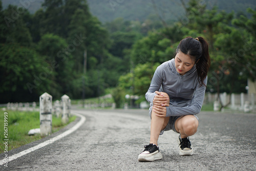 A young Asian woman crouching on the road holding her painful knee due to a joint injury during an outdoor running workout.