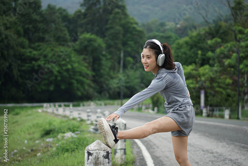 A medium shot of a cheerful Asian woman stretching her hamstring on a roadside post with green nature and mountains in the background.