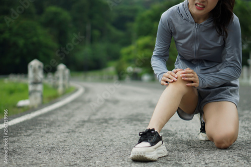Close-up photo of an Asian runner holding her knee in severe pain after suffering a joint injury on an asphalt road.