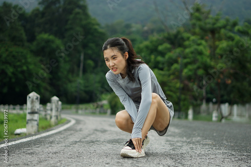 Full length photo of an Asian woman crouching on a road while holding her sprained ankle in pain during an outdoor workout.