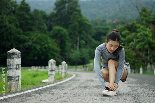 Full body shot of a young Asian woman looking down in discomfort while clutching her injured ankle on a scenic park road.