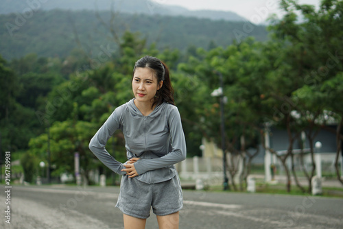 Medium shot of a young Asian woman holding her stomach in pain due to a muscle cramp during outdoor cardio exercise.