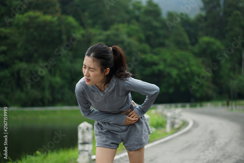 A young Asian woman clutching her side in severe abdominal pain and discomfort during an outdoor running exercise near a lake.