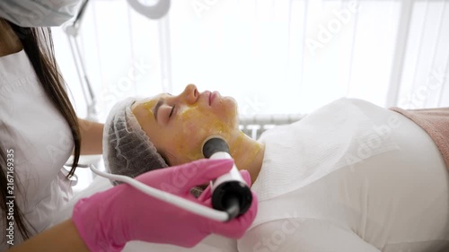 A woman receives a facial treatment with a handheld device applied to her face covered in yellow gel. The practitioner in pink gloves moves the tool across her skin in a spa setting