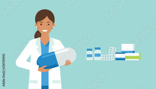 Friendly female pharmacist or doctor holding a giant pill capsule next to various pharmaceutical medications and pills.