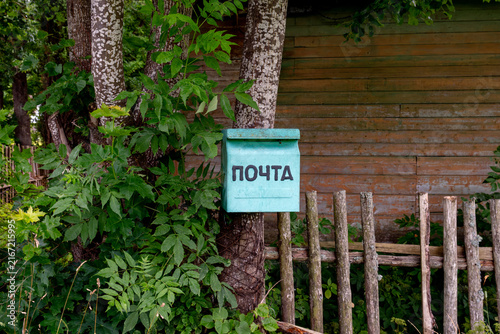 Izborsk Village, Pskov Oblast, Russia - July 26, 2026.A turquoise metal mailbox with the word mail written on it is attached to a tree trunk next to a rustic wooden fence and a weathered plank wall.