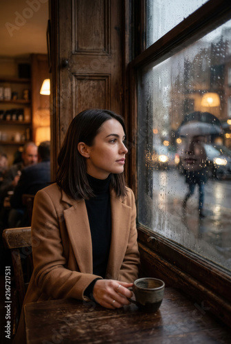 Woman Sitting in Cafe by Rainy Window with Warm Drink